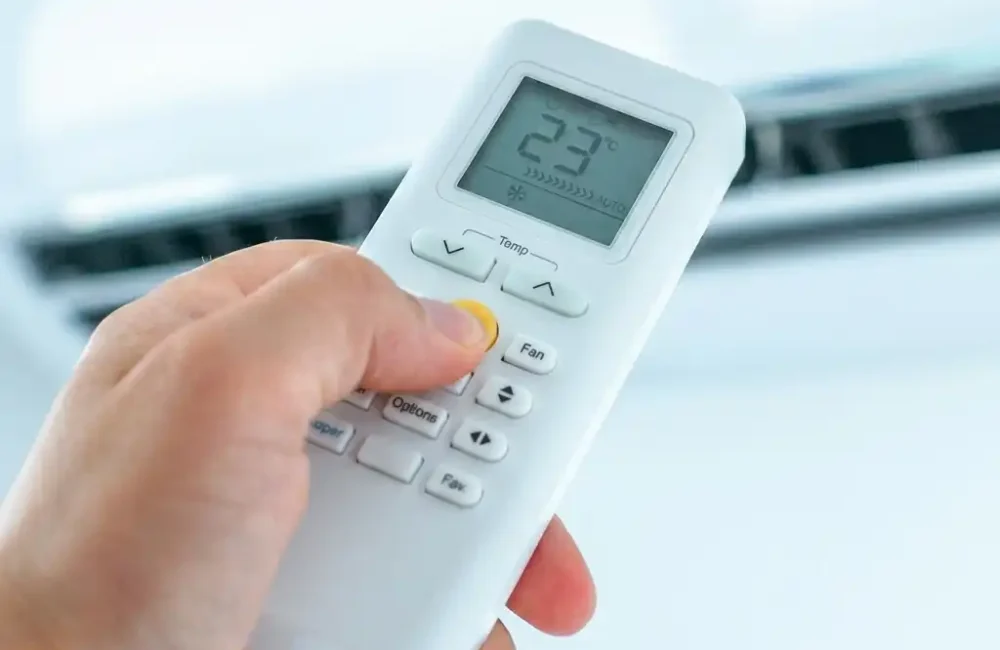 Main tenant une télécommande de climatisation réversible réglée à 23 °C devant une unité murale.