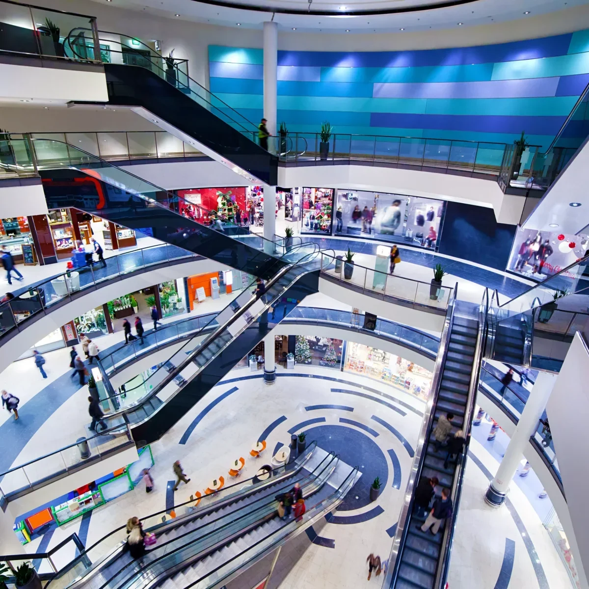 Vista interior de un centro comercial de varias plantas con escaleras mecánicas, tiendas y visitantes